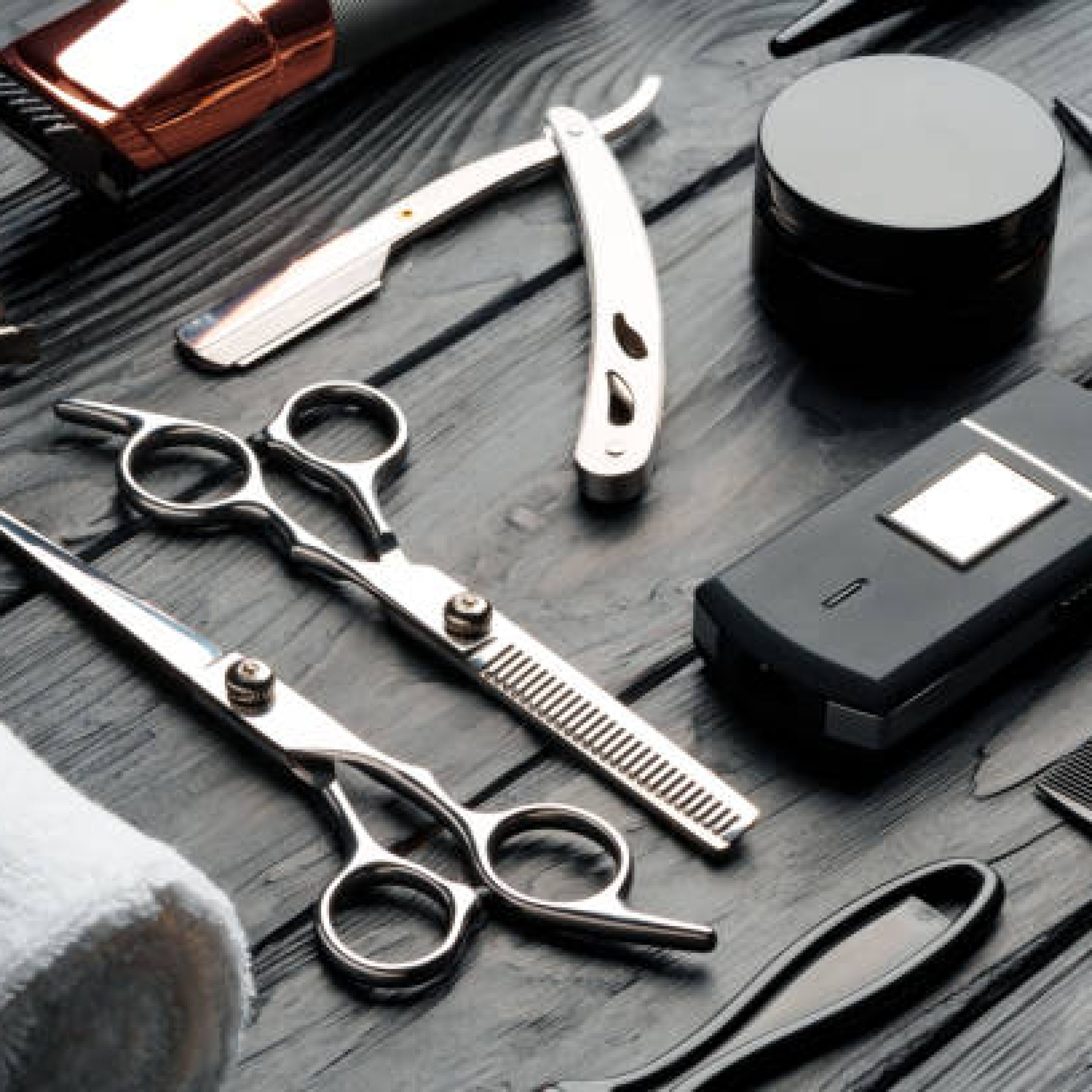 A variety of professional barber tools are neatly arranged on a dark wooden surface, including scissors, a straight razor, combs, clippers, and grooming products, suggesting preparation for a grooming session.
